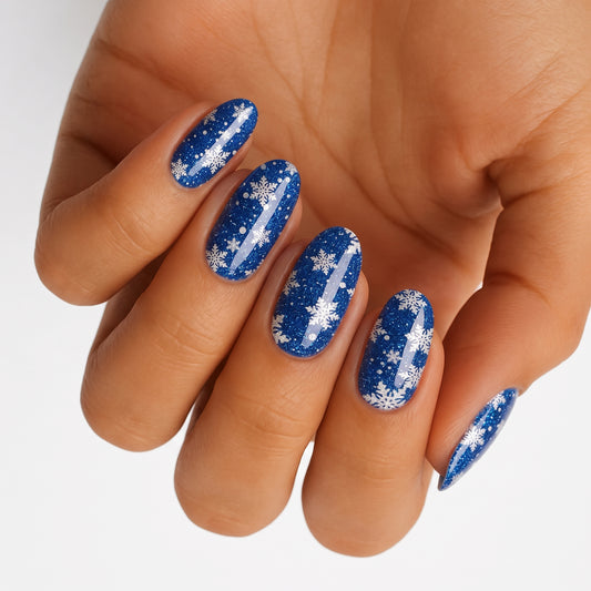 Glittery blue nail wraps with white snowflake designs on an oval shape